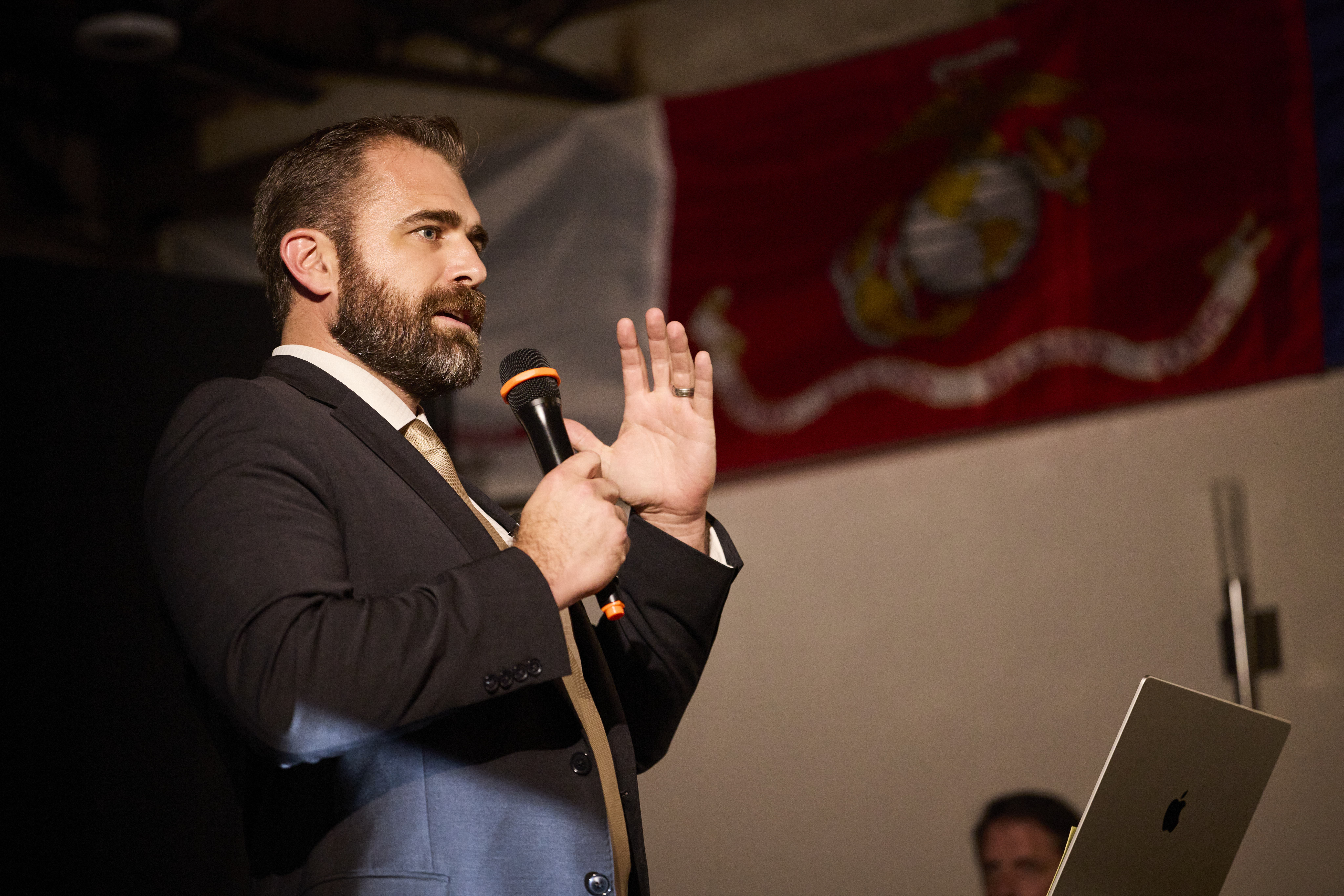 Dr. Chris Slininger delivering a keynote address, microphone in hand, in front of a U.S. Marine Corps flag